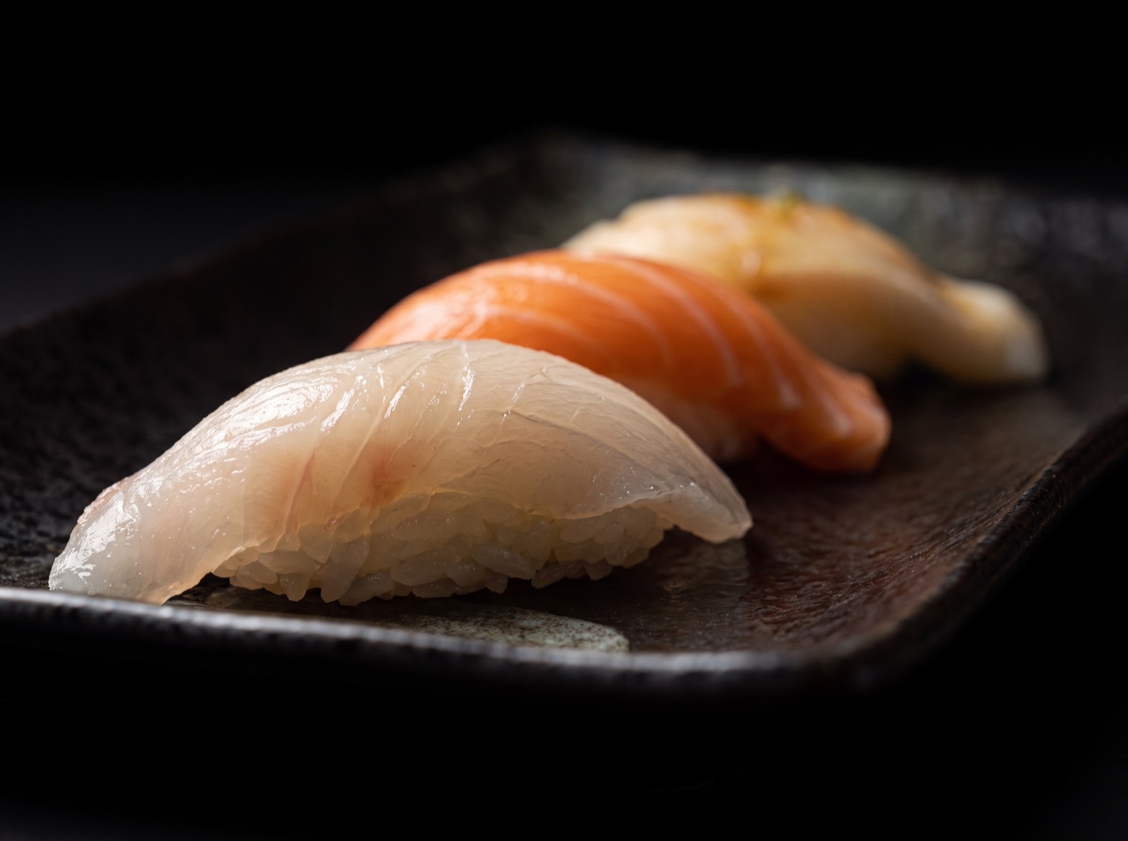 Salmon, white fish and yellowtail nigiri lined up on a dark slate plate with lemon.